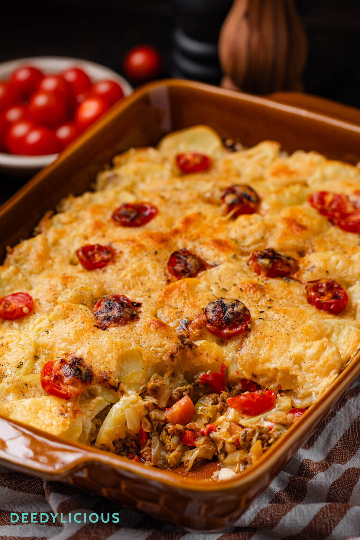 Oranje bruine ovenschaal met daarin de ovenschotel met spitskool, prei en komijn gemaakt door deedylicious. Daar omheen wat tomaatjes en specerijen die in het recept zijn gebruikt.