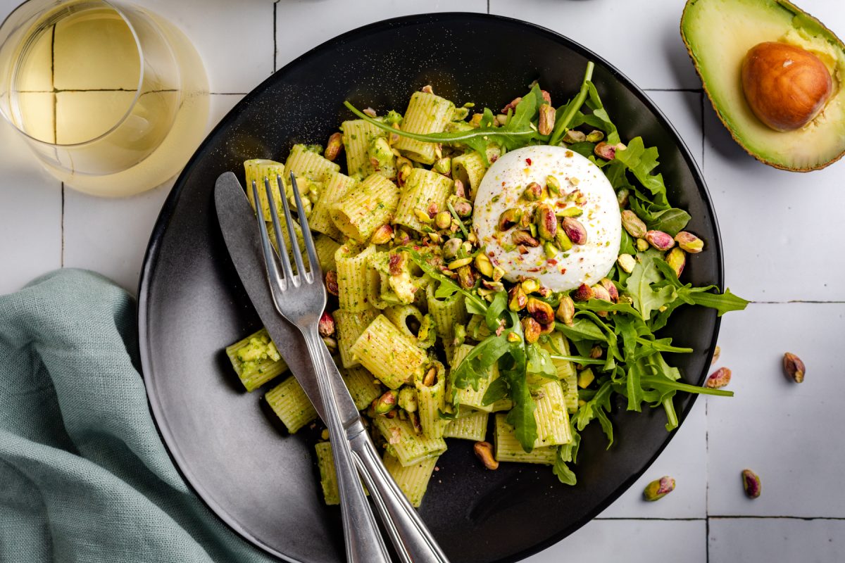 Rigatoni pasta met pesto, pistache en avocado, geserveerd op een zwart bord met witte tegels op de ondergrond