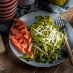 Een houten tafel met daarop een bordje groene past met daaroverheen parmezaanse kaas en wat coeur de boeuf tomaten. In de achtergrond staan een flesje Gran Moro balsamico, fles olijfolie een tomaat en een bosje basilicum.