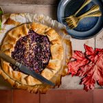 Een houten tafel met daarop een quiche met rode kool, daaromheen bordjes, herfstblad en een plantje basilicum.