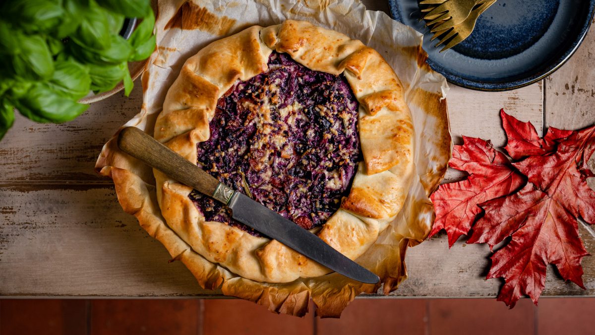 Een houten tafel met daarop een quiche met rode kool, daaromheen bordjes, herfstblad en een plantje basilicum.
