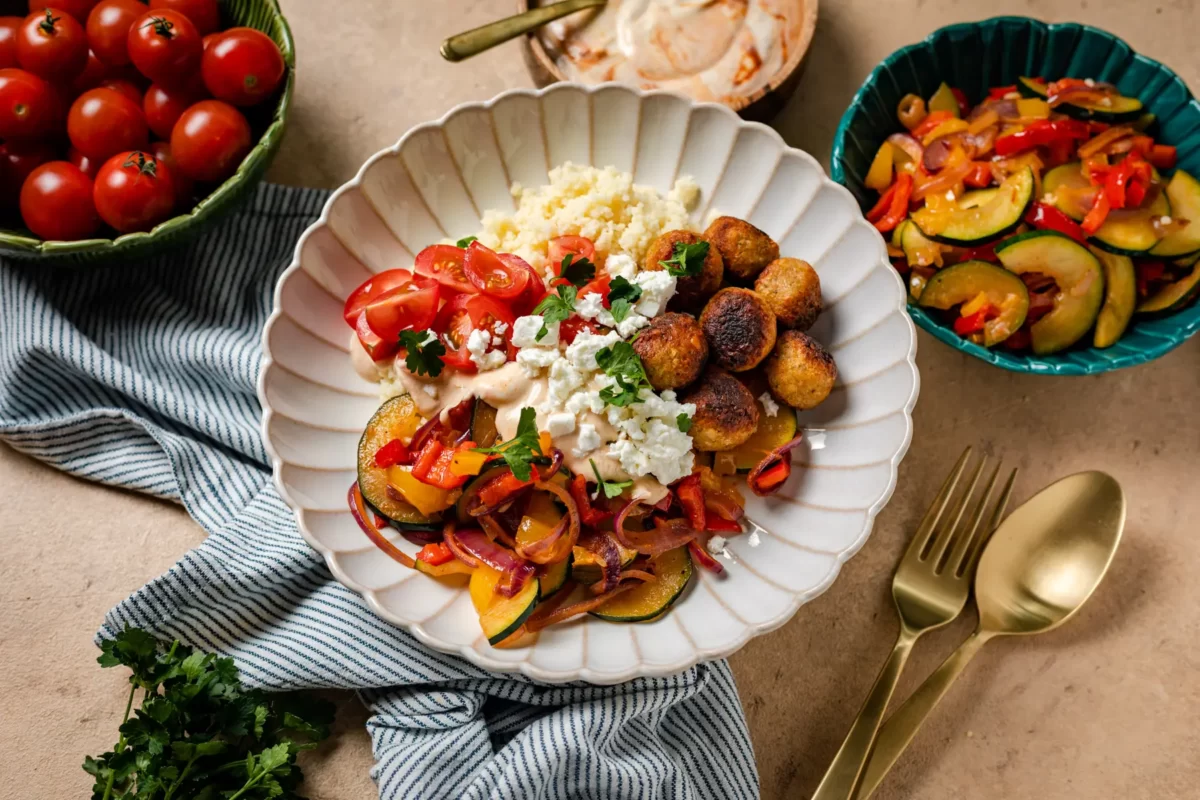 Falafel bowl op een geschulpt wit bordje met daaromheen goud bestek, schaaltje met tomaten, yoghurtdip met harissa en hummus, gegrilde groenten en peterselie. Onder het bord ligt een keukendoek.