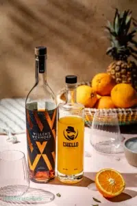 Een fles Chello arancello en Willem's Wermoed vermout met in de achtergrond een rieten mand met daarin sinaasappels en een ananas.