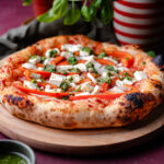 Een potje verse basilicum kruiden, een fles olijfolie rood en wit gestreept, een houten bord met daarop een vers gebakken pizza, een bakje met vers gemaakte pesto en in de achtergrond een rode backdrop met steenlook.