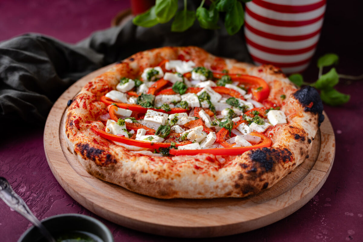Een potje verse basilicum kruiden, een fles olijfolie rood en wit gestreept, een houten bord met daarop een vers gebakken pizza, een bakje met vers gemaakte pesto en in de achtergrond een rode backdrop met steenlook.