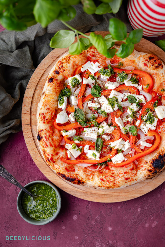 Flatlay met daarin een potje verse basilicum kruiden, een fles olijfolie rood en wit gestreept, een houten bord met daarop een vers gebakken pizza, een bakje met vers gemaakte pesto en in de achtergrond een rode backdrop met steenlook.