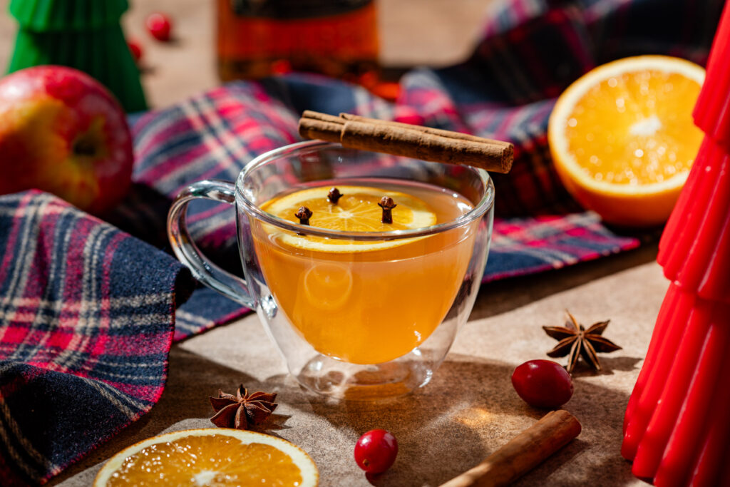 Warme appelsap met specerijen en spiced rum, in een dubbelwandig glas. Op de achtergrond zie je een tweede glas. In elk glas een schijfje sinaasappel met daarin kruidnagels gestoken.