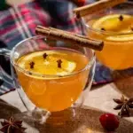 Warme appelsap met specerijen en spiced rum, in een dubbelwandig glas. Op de achtergrond zie je een tweede glas. In elk glas een schijfje sinaasappel met daarin kruidnagels gestoken.