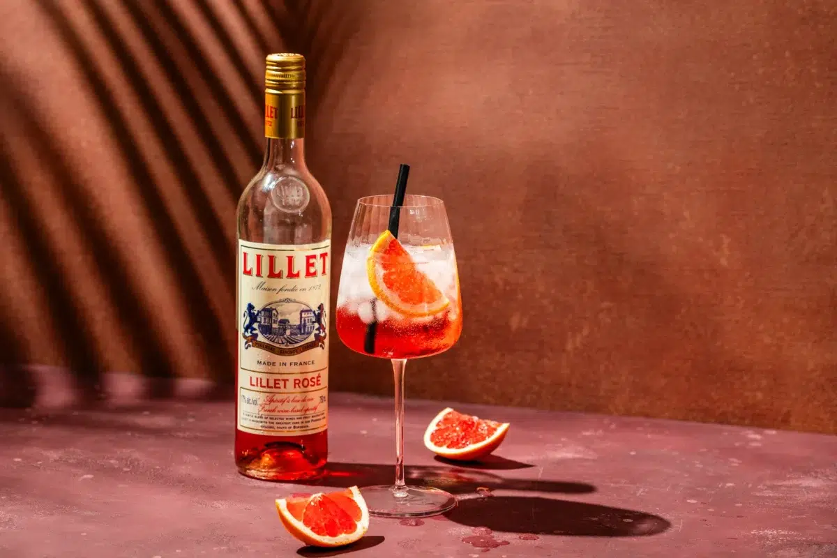 Lillet Rosé Tonic cocktail met daarachter een fles Lillet Rosé. Daaromheen liggen partjes grapefruit en in de achtergrond zie je een schaduw van een palmblad.