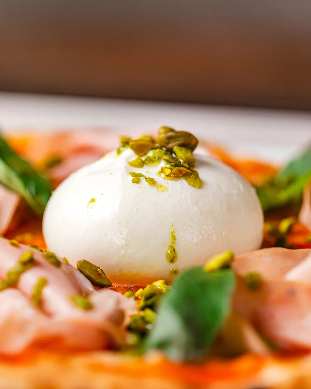 Detail van een pizza waar in het midden een bol mozzarella ligt. De mozzarella is groot in beeld en er liggen pistachenootjes op. Het is besprekende met basilicumolie en eromheen zie je nog net wat vleeswaren en blaadjes basilicum. Horeca fotografie door Wendy Pronk van DeedyLicious voor horecazaak in Leiden.