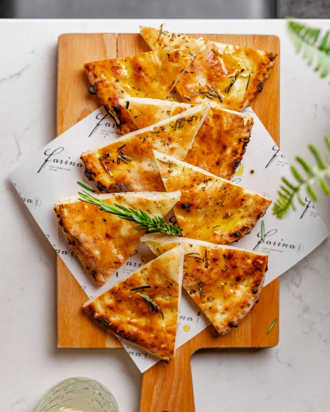 Een topshot van de bar van een pizzeria. Op de bar zie je een houten plank met daarop vetvrij papier waar een in punten gesneden focaccia ligt. Je ziet nog net een detail van een groene plant en een glas drinken in de hoek. Horeca fotografie door Wendy Pronk van DeedyLicious voor horecazaak in Leiden.