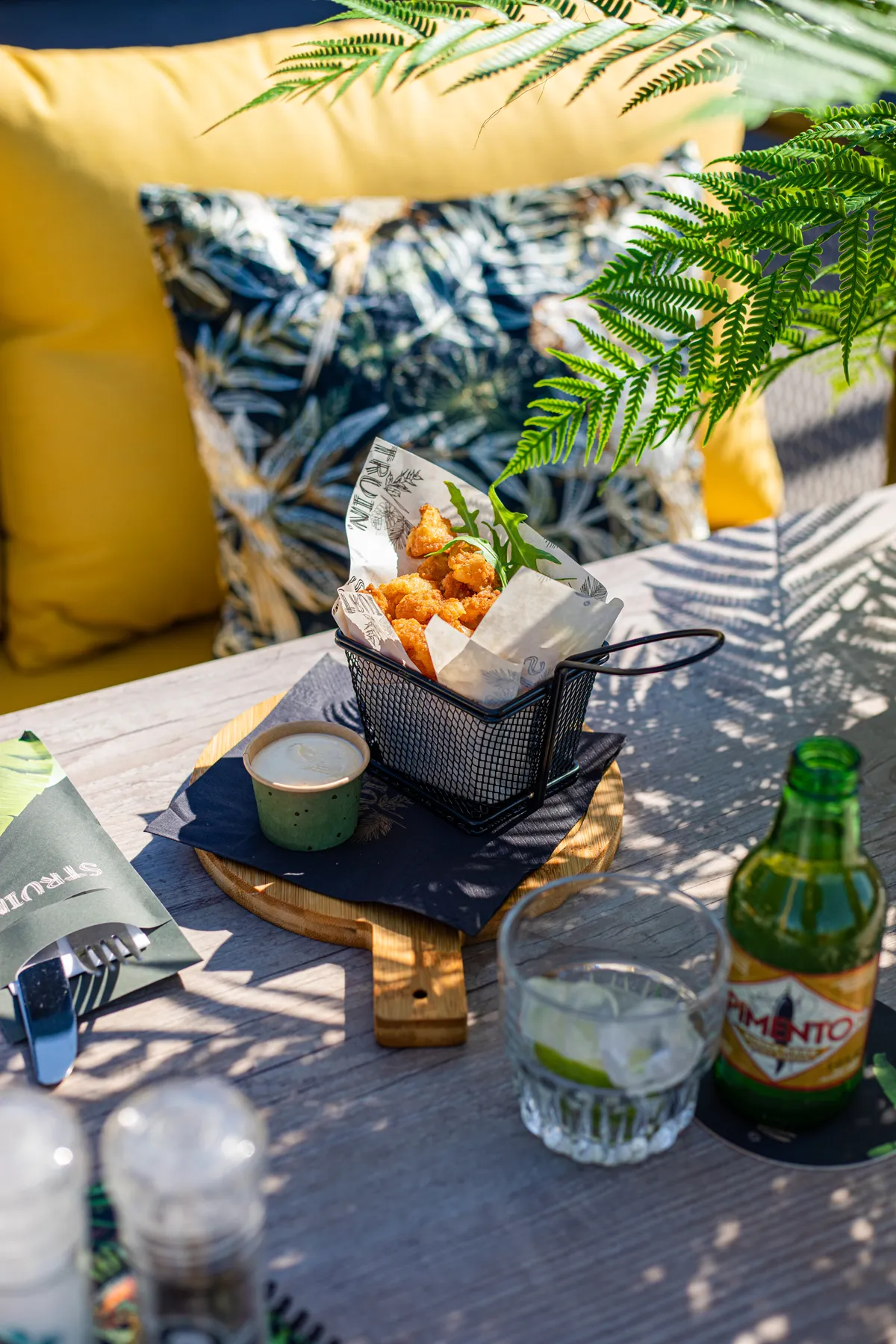Terrastafel met gele kussens in de achtergrond. Op de tafel staan diverse snacks en zie je de zon een schaduwspel spelen met de varen die op tafel staat.