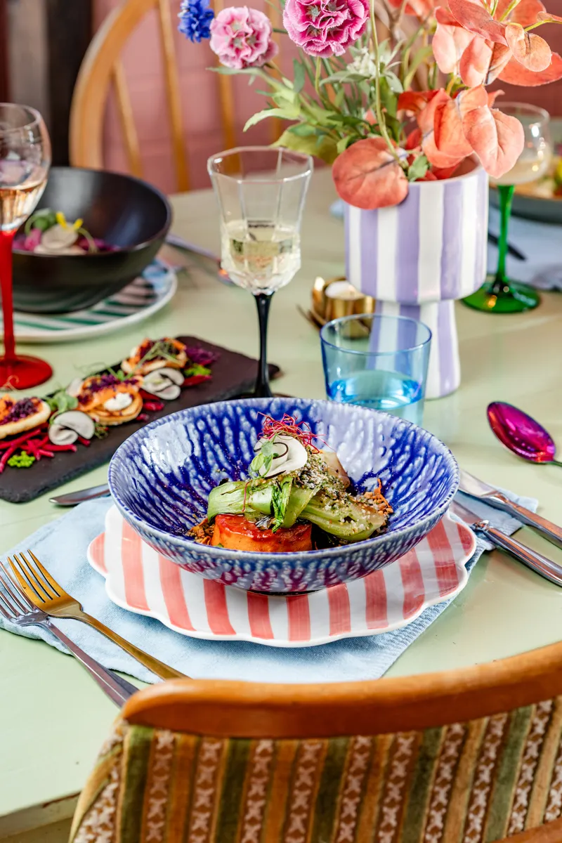 Gevulde eettafel bij vegan restaurant Ministerie van Geluk. Op tafel zie je een blauw bord met een kleurrijk gerecht erin. Daar omheen staan glazen wijn, een voorgerechtje en een kleurrijke bos bloemen.