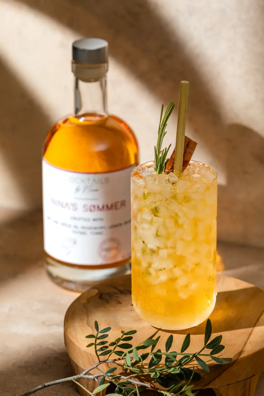 In een zomerse setting met warme beige tinten en de schaduw van planten in de zon, zie je een cocktail. Voor het glas ligt een takje groen en in de achtergrond staat de fles waar de cocktail uit komt.